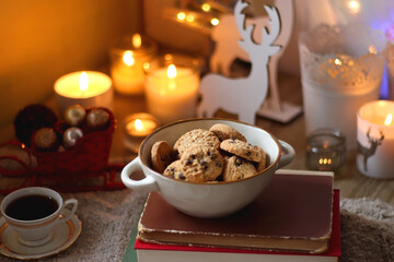 Bowl of cookies, cup of tea, dry oranges, pine cones, books, reading glasses, small presents, various Christmas decorations and lit candles on the table. Cozy Christmas hygge. Selective focus.