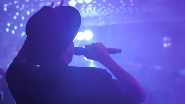 The reverse side of the silhouette of a man in the rays of spotlights and lanterns performs with a microphone on stage in front of a crowd of people. Live concert. singer. Slow motion