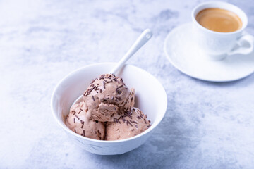 Chocolate ice cream with chocolate sprinkle in a white bowl on a light background. Three balls of homemade ice cream and a cup of coffee. Close-up.