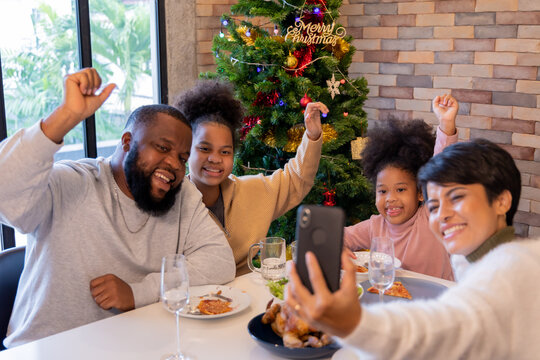 Cheerful african american family enjoying christmas and new years lunch while woman taking selfie on smartphone with members of family at home against decorated xmas tree