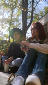 Two fashionable and spirited women, fully immersed in the hippie lifestyle, chat and enjoy their tea in the lovely autumn garden near the trailer.