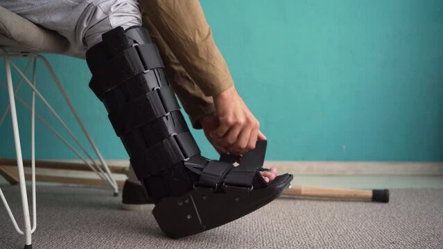 A man removing an ankle orthosis on a chair at home. Injured man in walker boots