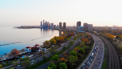 Top view of road, cars, dense tree crowns, train on rails, lake, skyscrapers. Cars drive along road to skyscrapers in distance. Concept enjoy evening cityscape on your road way home after hard day.