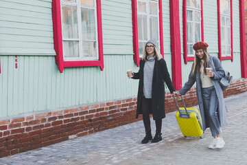 Two cheerful tourist women smiling and walking with suitcases on city street in autumn or spring time - travel and vacation concept