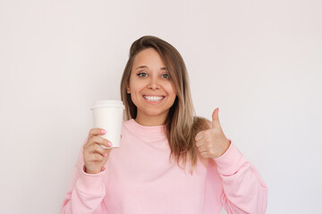 Young cheerful smiling blonde woman in a pink sweatshirt holding white eco paper cup with tea or coffee showing thumb up gesture isolated on a light background. Recycling eco-friendly concept