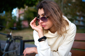 Portrait of a young woman in a modern sun glasses sitting on the wooden bench in a park near a lake enjoying the sunset.Stylish girl in light sports clothes sitting and relaxing on the beach at sunset