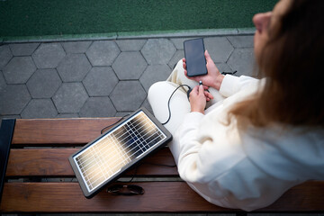 Young pretty woman sitting on the bench and charging her phone with a portable solar panel. Solar...