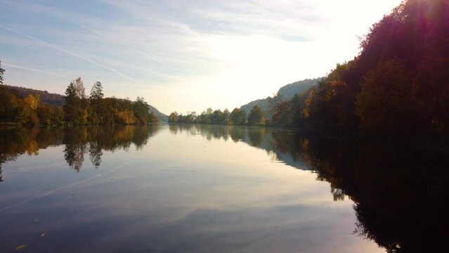 Drone flying low over river between colorful autumn leaf tree forest on sunny day