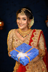 Portrait of a pretty young Indian woman dressed in traditional lehenga, gold jewellery and bangles holding gift box in hands on decorative background. Indian culture, occasion, religion and fashion.