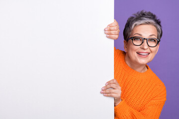 Photo of cute excited woman dressed orange sweater eyewear holding placard empty space isolated violet color background