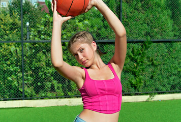 A cheerful attractive teenage girl holds a basketball in her hands above her head while standing on...