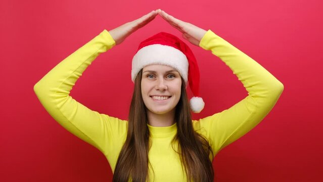 Merry young woman wear yellow sweater and Santa Claus hat hold folded hands above head like house roof, stay home, posing isolated over pastel red background wall. Happy New Year 2024 holiday concept