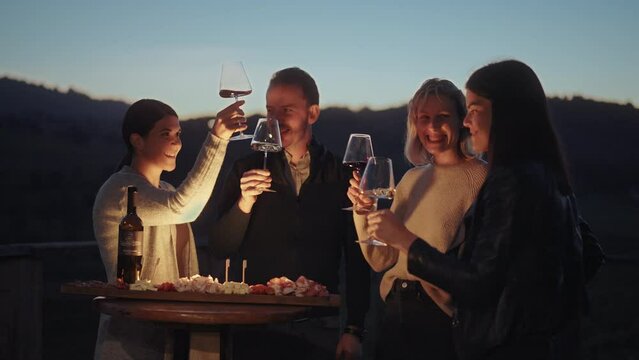 Group of friends enjoying wine tasting with local cheese, prosciutto and wine at sunset. Countryside vineyards and mountains in the background. Happy adult friends having fun, drinking wine and eating