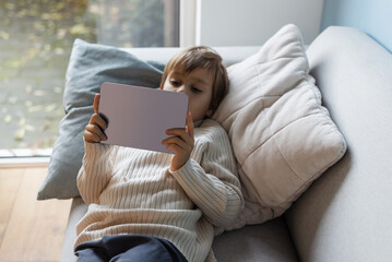 Little cute boy watching cartoons and playing games on a digital tablet lying on the sofa. - obrazy, fototapety, plakaty