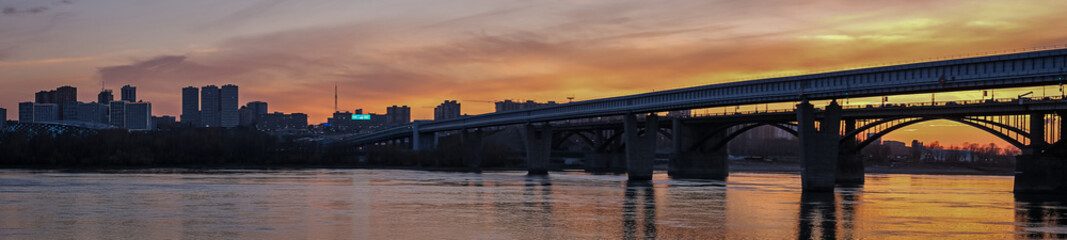 Fototapeta na wymiar Beautiful view of the bridge over which cars drive at sunset. A river flows under the bridge, reflecting the sunset rays. Bridge in the city of Novosibirsk, Russia