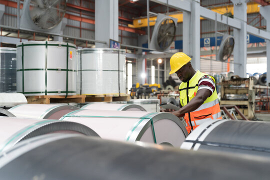 Metalwork manufacturing, warehouse of raw materials. Male factory worker inspecting quality rolls of metal sheet in aluminum material warehouse, wearing safety uniform and uses digital tablet - Powered by Adobe