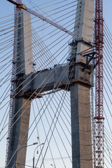 Fototapeta premium Crane and bridge construction against a blue sky background. Builders work on large construction sites, and there are many cranes working. There are a lot of cables on the bridge