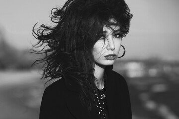 High fashion concept. Gorgeous young girl with long dark glossy hair in black dress and coat on the beach. Cinema star, diva style. Close up. Text space. Monochrome outdoor shot