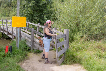 Mature hiker together with his dog opening and crossing gate towards bridge leading to nature...
