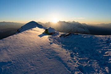 Winterbiwak im Schnee auf einem Berg in den Alpen mit grünem Zelt zum Sonnenaufgang und Blick auf...