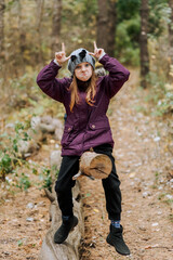 Beautiful smiling happy girl, child makes faces, grimaces, shows horns, sitting on a log in the forest in autumn. Photography, portrait.