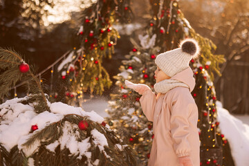 A little girl in winter clothes has fun throwing snow while standing on a decorated Christmas street in winter clothes. Christmas or New Year greetings concept. 