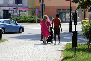 Kobieta z wózkiem i mężczyzna o ciemnym kolorze skóry idą chodnikiem.