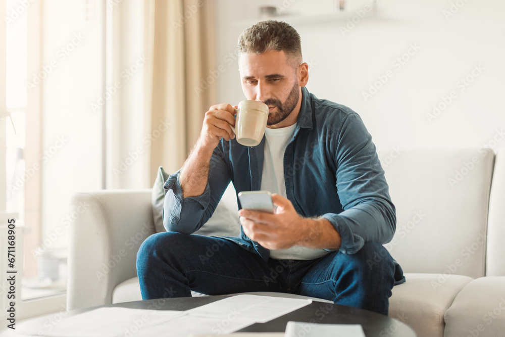 Poster man using smartphone while managing paperwork and drinking coffee indoor