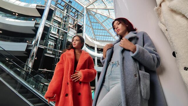 Two trendy woman high fashion model in stylish fur coat posing at modern mall shopping center outlet