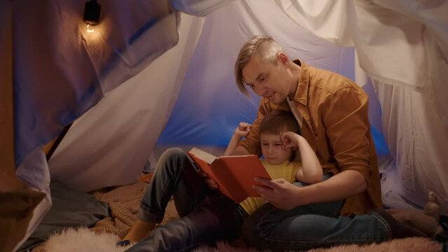 A dad along with his son reading a book, in a tent in the living room. The son is lying with his back resting on dad's chest, and points his thumb up when dad points to the book pages.