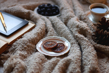 Bowl of cookies, cup of tea or coffee, plate of blueberries, dry oranges, wool yarn, pine cones, books, reading glasses, tablet, pen, soft blanket and lit candles on the table. Winter hygge at home.