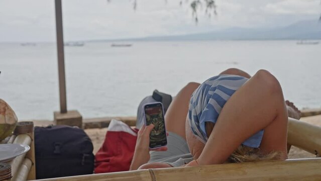  Young woman in a blue Headscarf lies on the beach, on the ocean shore, and looks at pictures on her phone. Slow Motion
