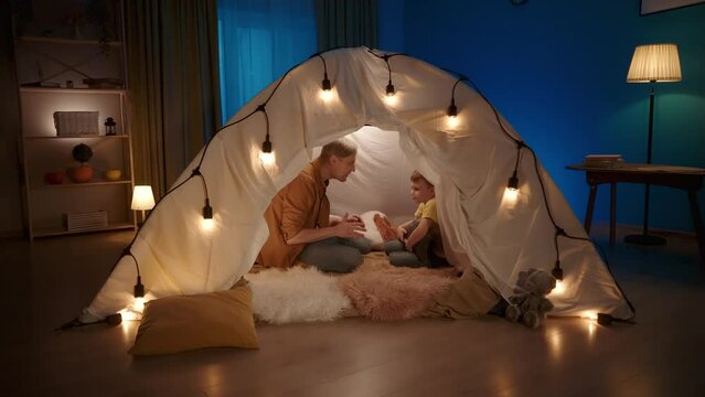 Dad and son's night out together. A man and a boy sitting in a garland lit tent in a dark living room. Dad tells scary fantasy stories, tickles the child and bites him. Concept of happy childhood.