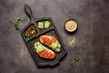 Open sandwich with salmon, cucumber and microgreens on a wooden cutting board, dark rustic background. Top view, flat lay, copy space.