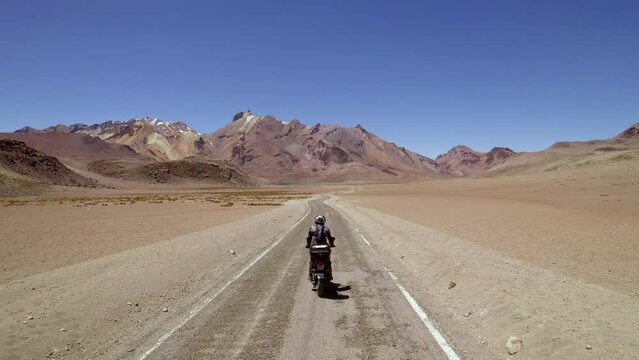 man on motorcycle closely followed at high speed by drone on lonely desert road with hills and lagoons 