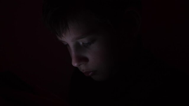 Cute little boy sitting in the dark and watching cartoons on mobile. Smiling toddler laying on bed and looking at smartphone. Light reflection on kid face from phone.