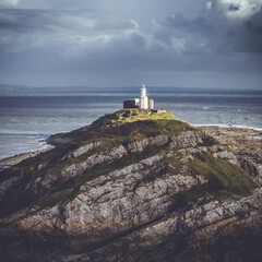 Fototapeta na wymiar Experience the captivating allure of Mumbles Lighthouse in this vibrant square format photograph. Against the dramatic backdrop of a stormy sky, the lighthouse stands tall, bathed in the soft glow of 