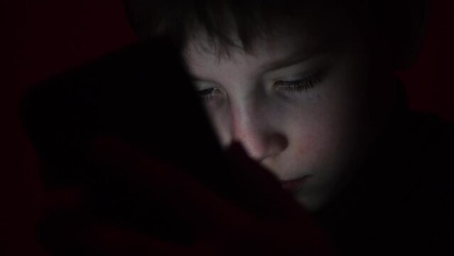Cute little boy sitting in the dark and watching cartoons on mobile. Smiling toddler laying on bed and looking at smartphone. Light reflection on kid face from phone.