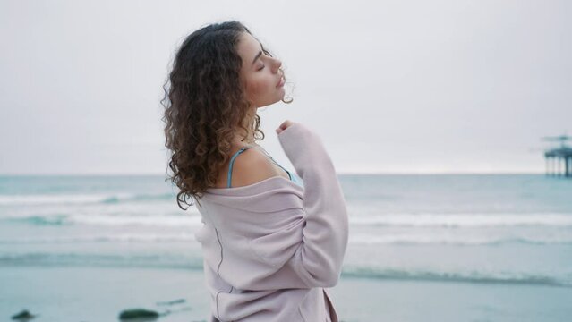 Fashion model RED camera. Gorgeous girl poses at ocean, emotionally looking at camera. Portrait of beautiful curly woman with natural makeup in shiny metallic sheer blouse at beach on gloomy grey day