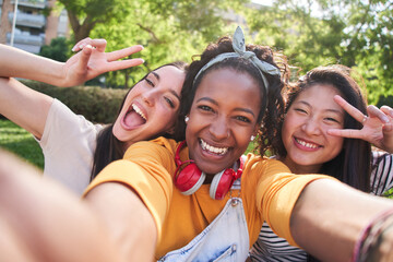 Selfie three excited multicultural cheerful young women outdoors. Females having fun looking at smiling camera and making peace symbol with hands enjoying summer vacation. Generation z in sunny park. - Powered by Adobe
