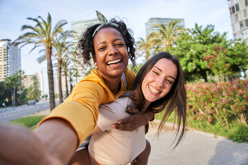 Multiracial lesbian young couple taking selfie portrait outdoor sunny day. Beautiful Caucasian woman giving piggyback ride to nice African girl on city street. Smiling female posing for photo happy.