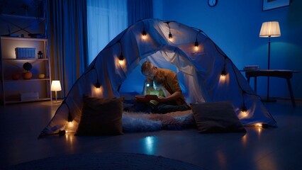 Dad and son, are reading a book, in a tent in a dark living room. The son is sitting with his back resting on dad's chest, holding a switched on flashlight directed at the pages of the book. - obrazy, fototapety, plakaty