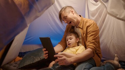 Father and son taking selfies with a tablet, making funny faces, close up. Father and son having fun in a camping tent decorated with garlands, in their living room in the evening.