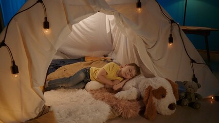 A boy sleeping on a big soft toy, a dog, instead of a pillow, on fur plaids in a tent, which is set...