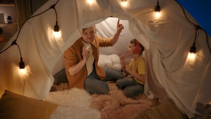 Side view of father and son wearing wireless headphones listening to music and dancing. Man and boy sitting in garland lit tent in dark living room. Close up. A father and son's evening together. - obrazy, fototapety, plakaty