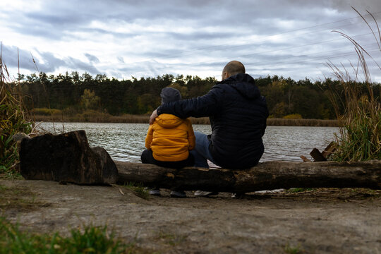 A father hugs his son while sitting on the shore of a lake. Father and son sitting on a fallen tree near the lake