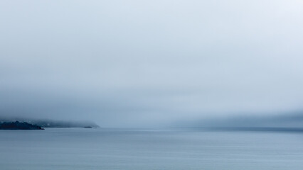 Une atmosphère mystérieuse en Bretagne avec une épaisse brume, des ciels gris, et la côte bretonne visible à l'horizon. La mer au premier plan est remarquablement calme, reflétant des tons gris et ble