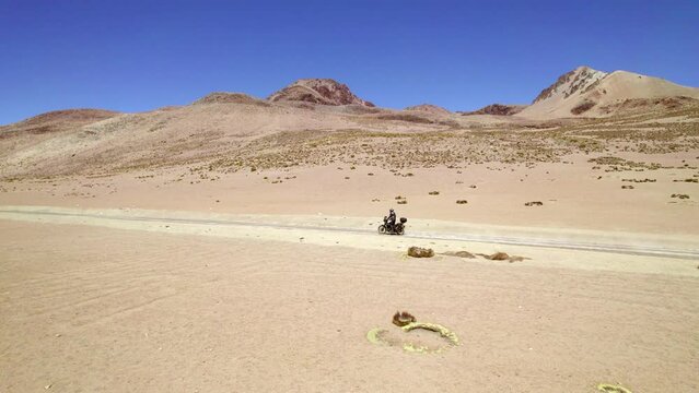 side view man on motorcycle followed by drone discovering a lonely desert road with mountains and lagoons 