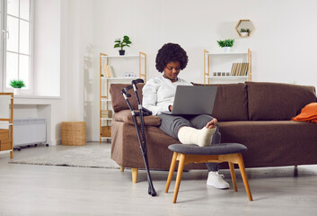 African American woman with fractured leg sitting on sofa and using laptop computer. Beautiful girl with plastered leg sitting on couch working online while recovering at home - obrazy, fototapety, plakaty