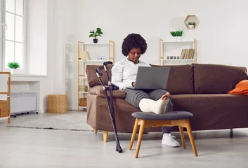 Fotobehang African American woman with fractured leg sitting on sofa and using laptop computer. Beautiful girl with plastered leg sitting on couch working online while recovering at home © Studio Romantic